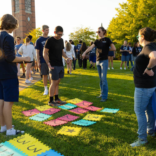 on Kirkhof Center lawn, student steps on plastic squares that are numbered for a math game, a table with Department of Mathematics is nearby, two faculty members standing
